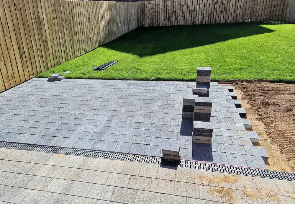 A backyard patio made of gray paving stones is partially completed by a construction company in Morris & Essex County, NJ. Stacks of unused pavers remain on top, with a green lawn and a wooden fence in the background.