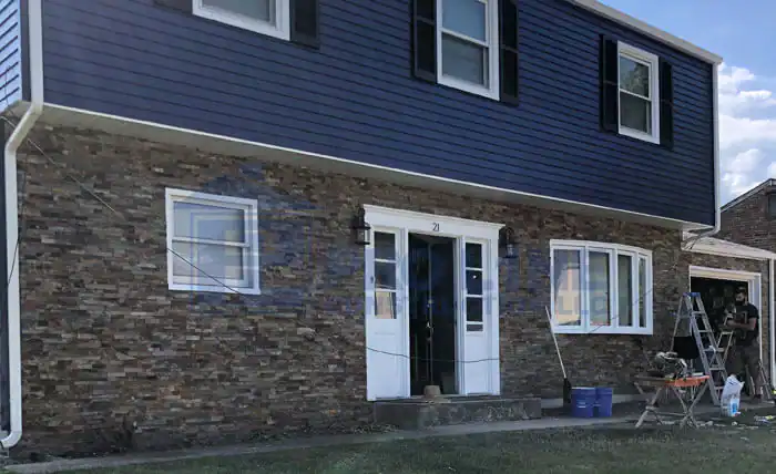A two-story house with blue siding and stone brick on the lower level, white-framed windows, black shutters, and a white front door. Workers from a construction company in Morris & Essex County, NJ are busy near the open garage on the right.