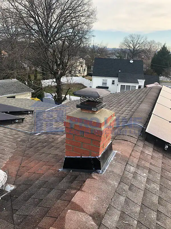 A brick chimney with a metal cap extends from a shingled roof. Solar panels, installed by a leading construction company in Morris & Essex County, line the roof edge, with trees and neighboring NJ houses in the background.