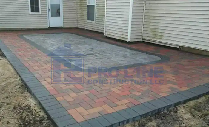 A rectangular backyard patio made of multi-colored bricks with a decorative border and inner pattern, designed by a leading construction company in Morris & Essex County, NJ, adjacent to a tan house with siding and a glass door.
