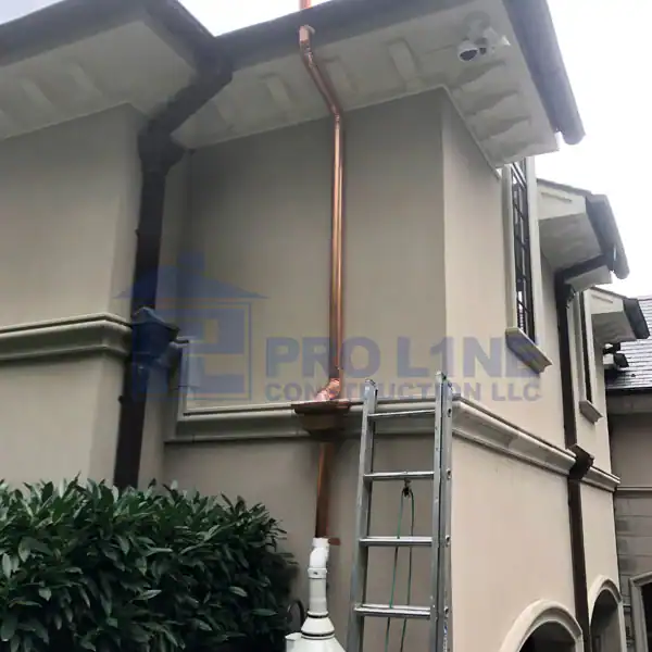 A copper downspout is installed on the side of a two-story beige house with white trim by a construction company in Morris & Essex County, NJ. A ladder leans against the wall, and bushes are visible in the foreground. A security camera is mounted under the eaves.