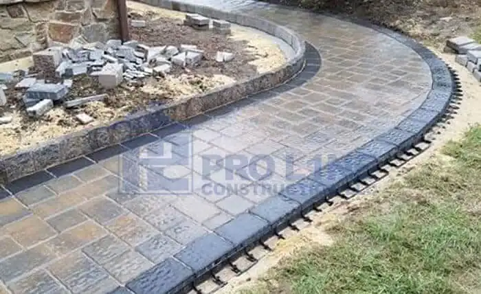 A curved stone walkway with black brick edging is partially completed by a trusted NJ construction company in Morris & Essex County, with loose bricks and sand on one side and grass on the other.