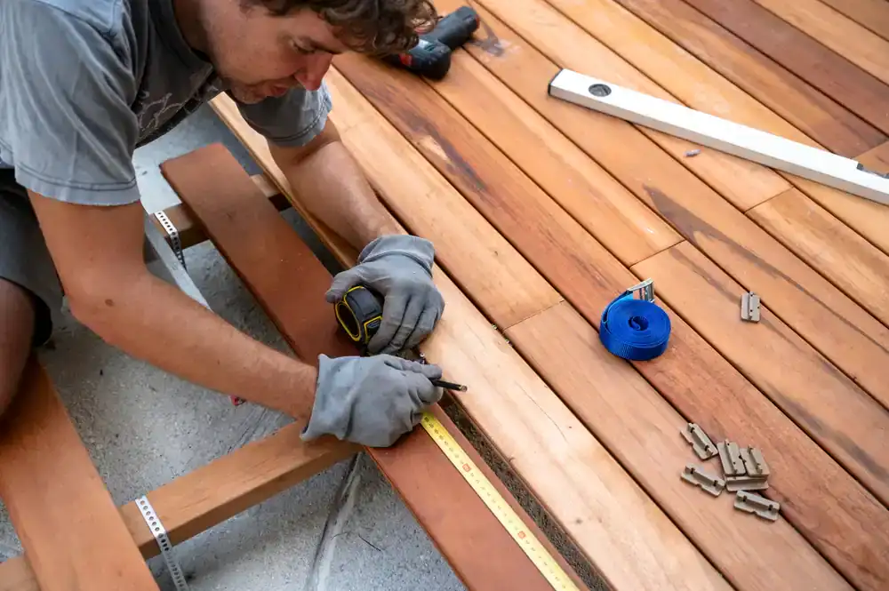 A person wearing gloves measures and marks a wooden plank with a tape measure and pencil, preparing to install it among other boards&mdash;a typical scene for a construction company in Morris & Essex County, NJ. Tools and wooden spacers are scattered around.