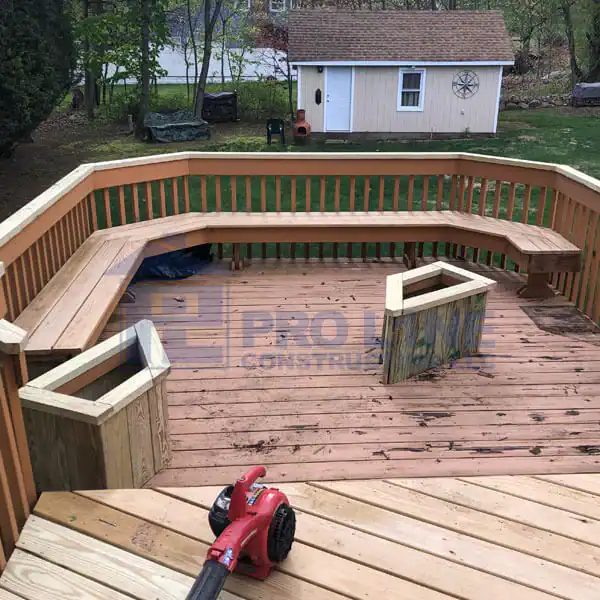 A wooden backyard deck by NJ construction company Morris & Essex County features built-in benches, two square planter boxes, and scattered leaves. A red leaf blower sits on the decking, with a shed and yard visible in the background.