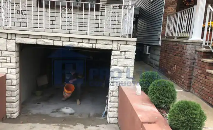 A person crouches inside a small garage or basement with stone walls, holding an orange bucket. The driveway slopes down near green bushes and brick steps—typical of homes serviced by an NJ construction company in Morris & Essex County.