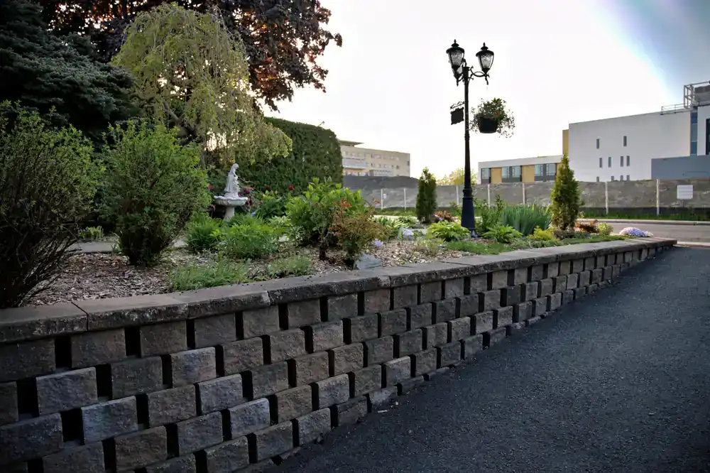 A raised stone garden bed with various green plants and flowers, a small statue fountain, and a vintage-style streetlamp&mdash;outdoor charm designed by an NJ construction company in Morris & Essex County. Modern buildings and a paved path are visible in the background.
