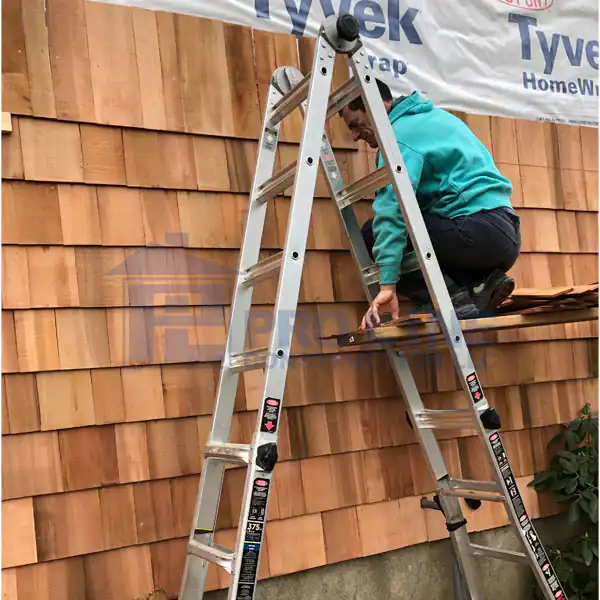A person in a teal hoodie is kneeling on a folded ladder while working on a house exterior with cedar shingles and Tyvek HomeWrap—an unsafe position not recommended by any reputable construction company in Morris & Essex County, NJ.