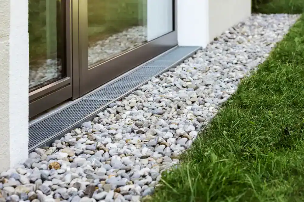 Gray pebbles line the ground next to a building with a glass door, bordered by a metal drainage grate and a strip of green grass&mdash;typical of landscaping by an NJ construction company in Morris & Essex County.