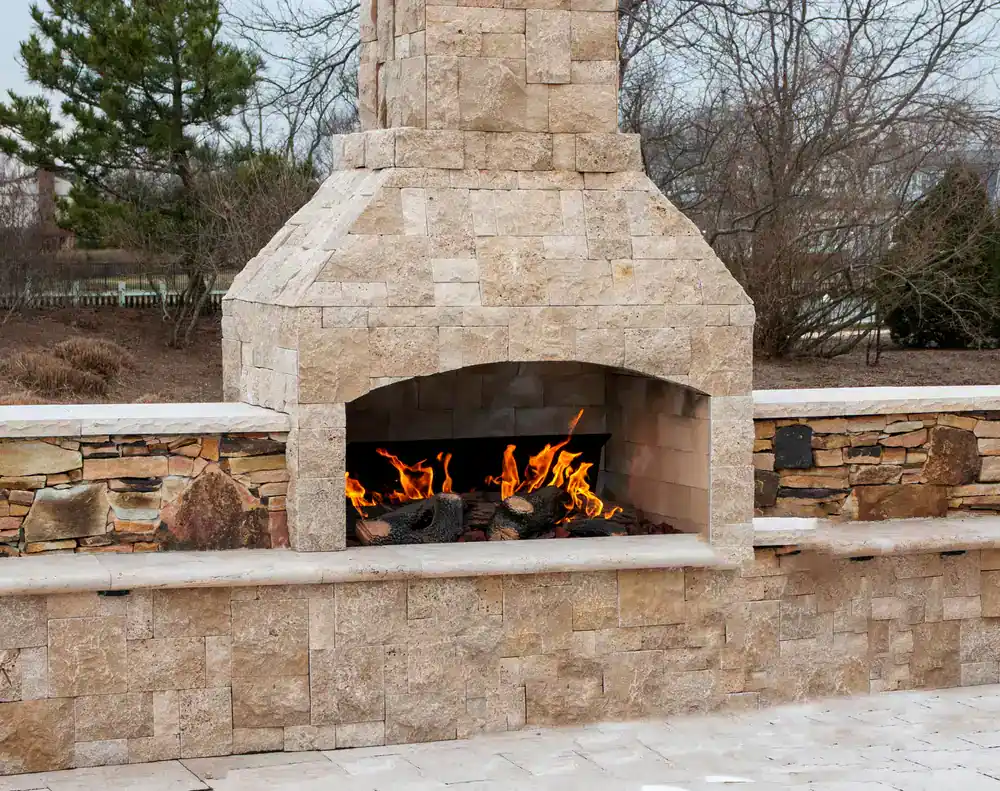 Outdoor stone fireplace with a burning fire, surrounded by a stone patio and wall. Designed by a trusted construction company in Morris & Essex County, NJ. Leafless trees and a cloudy sky are visible in the background.