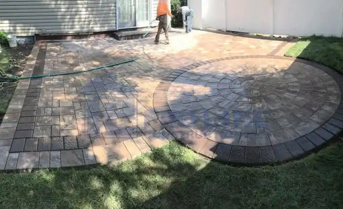 A backyard patio with decorative stamped concrete in rectangular and circular patterns. Two people from a construction company in Morris & Essex County, NJ are working, and the area is partly shaded, with grass bordering the patio.