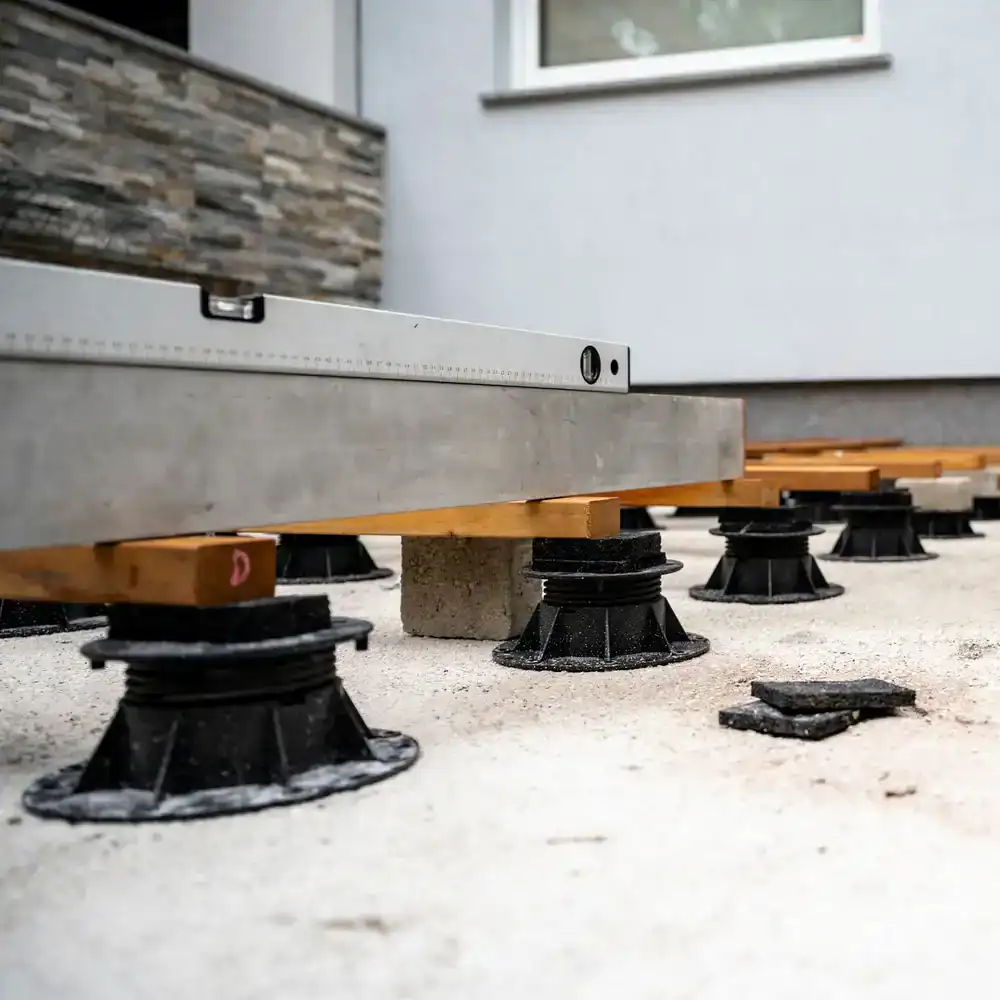 Close-up of adjustable black deck supports on a concrete surface, with wooden beams and a metal level tool placed on top, near a modern building exterior&mdash;ideal for any construction company in Morris & Essex County, NJ.