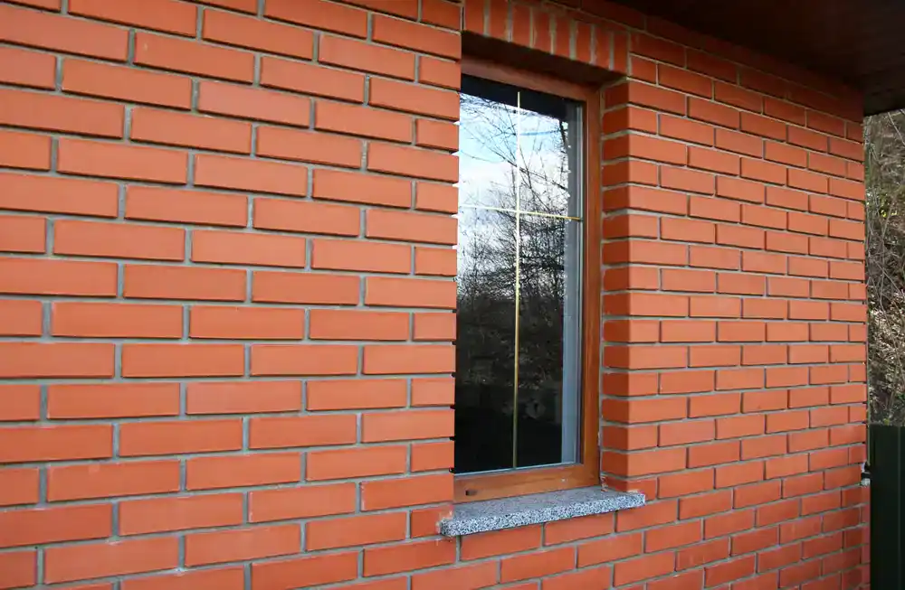 A close-up of a red brick wall with a rectangular window featuring a wooden frame and glass panes, reflecting trees and sky&mdash;a signature touch often seen in projects by construction company Morris & Essex County, NJ.