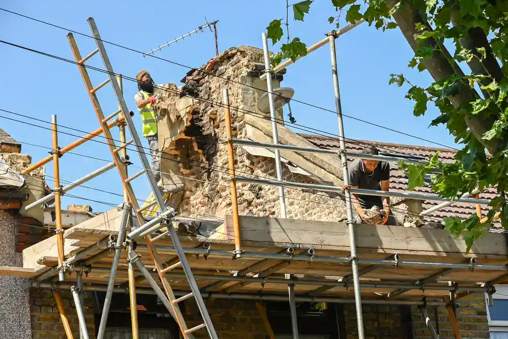 Two construction workers from an NJ construction company repair a damaged brick chimney on a house roof. One works near the crumbling structure while the other stands nearby. Trees and power lines are visible in the Morris & Essex County area foreground.