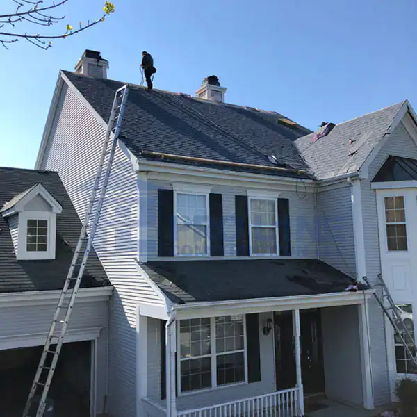 A person stands on the roof of a two-story house, apparently working on repairs. Several ladders from a construction company in Morris & Essex County, NJ, are propped against the house beneath a clear blue sky.