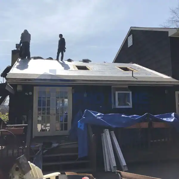 Two workers stand on the roof of a house under construction or repair in Morris & Essex County, NJ. The roof shows new underlayment, while construction materials and equipment from a local company are visible around the house and yard.