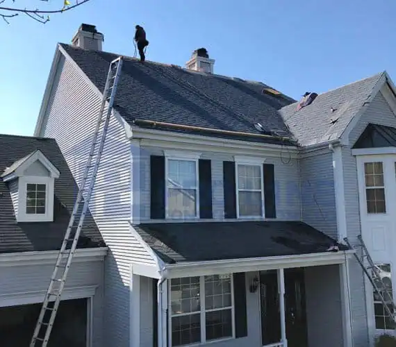 A person stands on the steep roof of a two-story house in Morris & Essex County, NJ, with two tall ladders leaning against the house. The roof appears under repair by a construction company, with some shingles missing and a clear blue sky above.