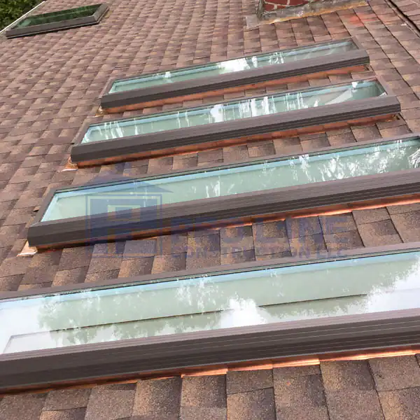 Four rectangular skylights are installed in a row on a shingled brown roof by a leading construction company in Morris & Essex County, NJ, allowing natural light into the building below. Trees are reflected in the glass.