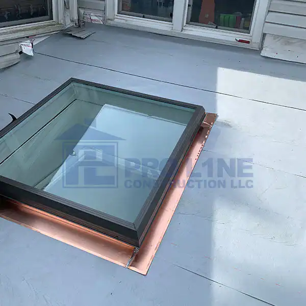 A glass skylight with a black frame and copper flashing is installed on a light gray flat rooftop by a leading construction company in Morris & Essex County, NJ, near a building with white siding and windows.