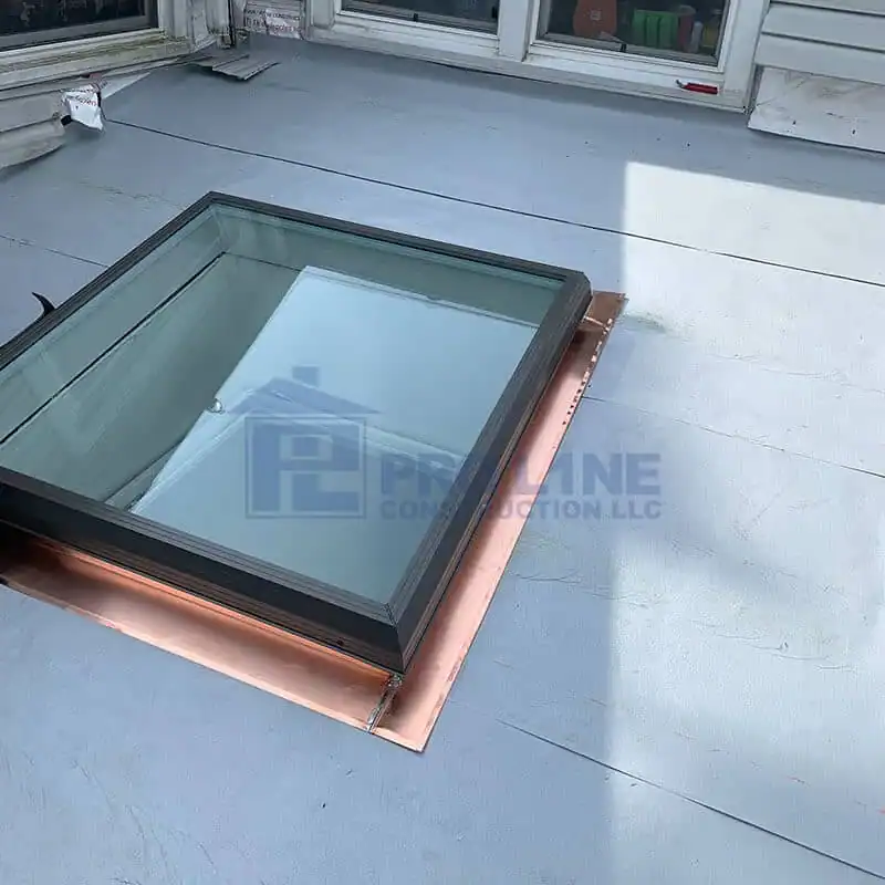 A skylight window with a black frame is installed on a flat gray rooftop by an NJ construction company in Morris & Essex County, surrounded by copper flashing. The roof surface appears clean and smooth.