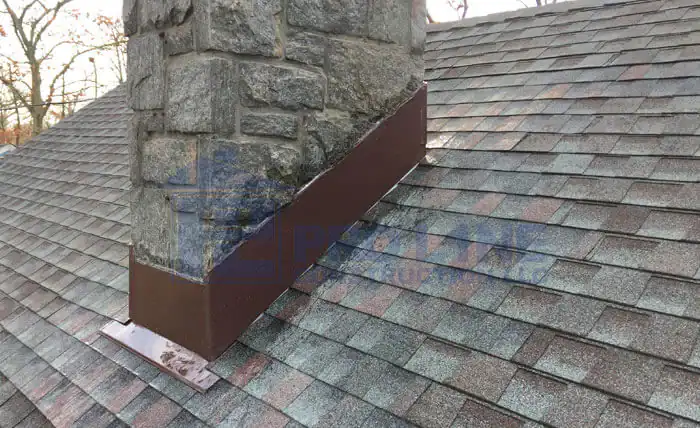 A stone chimney with brown metal flashing is installed on a shingled sloped roof by a trusted NJ construction company; the flashing seals the base of the chimney to prevent water leaks.