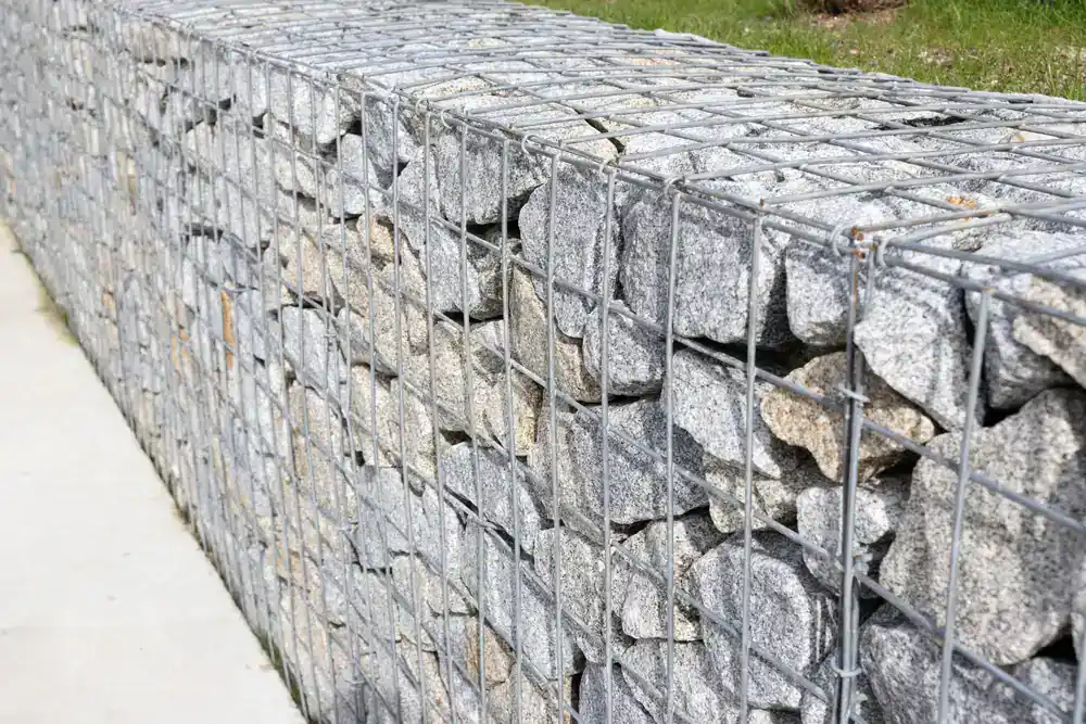 A close-up view of a gabion wall, built by an NJ construction company in Morris & Essex County, features gray rocks held by metal wire mesh, with grass visible in the background.