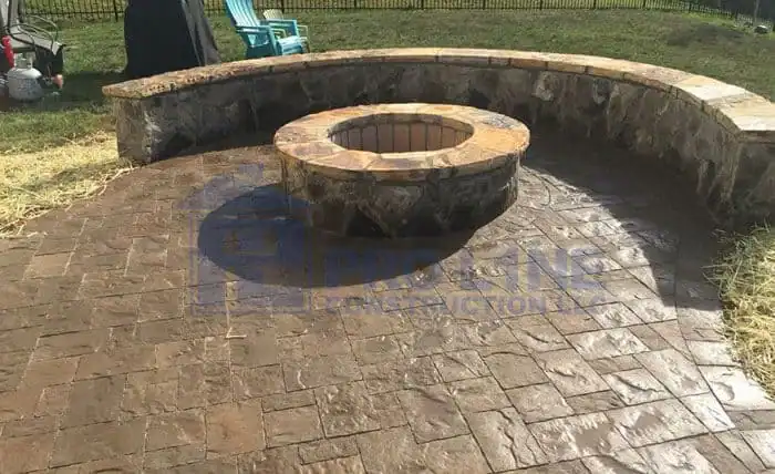 A circular stone fire pit with a matching curved stone bench sits on a stamped concrete patio in a backyard, crafted by an expert construction company in Morris & Essex County, NJ. Two blue chairs and some straw are visible in the background.