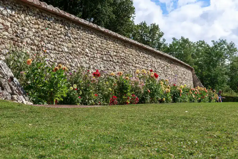 A stone wall borders a lush green lawn, with colorful flowers including red, yellow, and pink blooms planted along the base. Created by a top NJ construction company in Morris & Essex County, trees and a partly cloudy sky complete the scene.