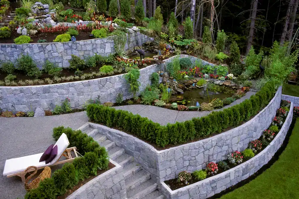 A multi-level terraced garden, crafted by a top construction company in Morris & Essex County, NJ, features stone retaining walls, vibrant flowers, shrubs, and a small pond. Steps connect each level to a lounge chair on the lowest terrace, surrounded by greenery.
