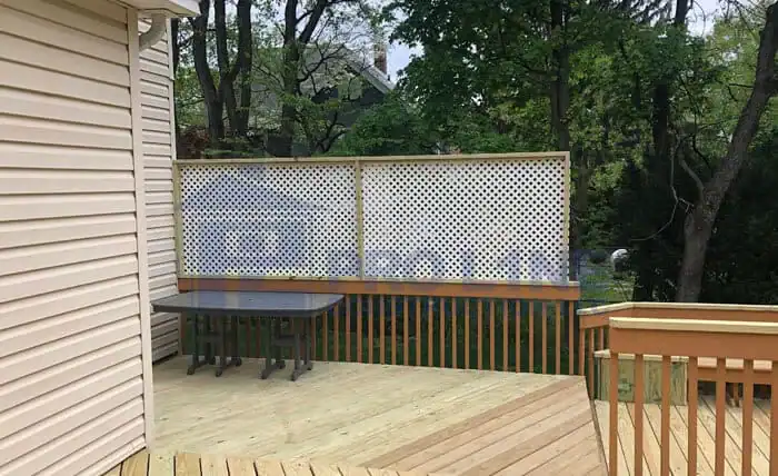 A wooden deck with a table, surrounded by a wooden railing and a lattice privacy screen, crafted by a skilled construction company in Morris & Essex County, NJ. Trees and houses are visible in the background.
