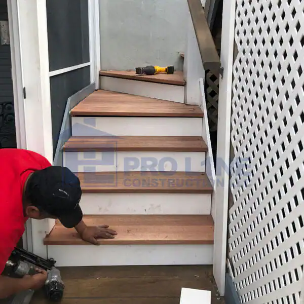 A person in a red shirt installs wooden steps on a small staircase for a construction company in Morris & Essex County, NJ, using a tool. A drill sits on the top step, with a white lattice panel visible on the right side.