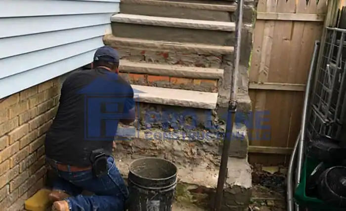 A worker from a construction company in Morris & Essex County, NJ, kneels on the ground, repairing concrete steps beside a brick wall and wooden fence. A bucket and various tools are nearby, and the steps appear to be freshly patched.