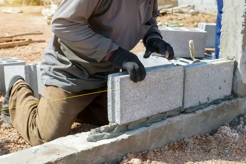 A construction worker wearing gloves kneels on the ground, positioning concrete blocks and using a yellow guideline to ensure alignment while building a wall for an NJ construction company in Morris & Essex County.