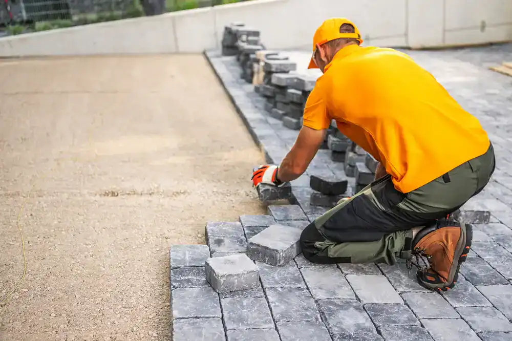 A worker in orange clothing kneels on the ground, laying gray concrete pavers for a walkway or patio&mdash;typical of expert projects by a construction company in Morris & Essex County, NJ. Stacks of unused pavers are nearby.
