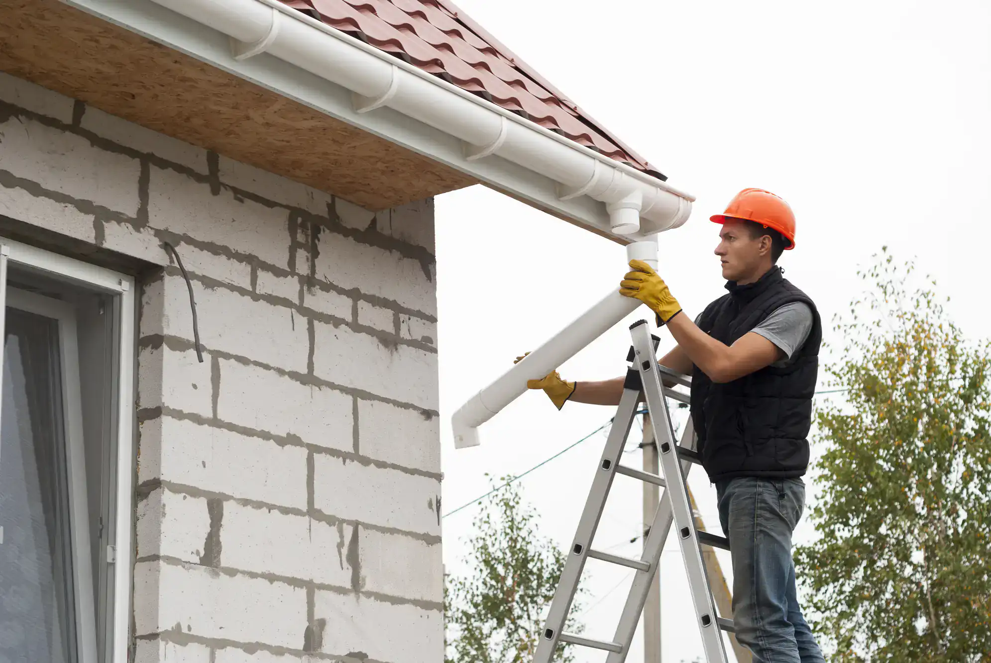 A construction worker wearing a hard hat and gloves stands on a ladder, installing a white rain gutter on the roof edge of a brick house under construction. Trees are visible in the background.