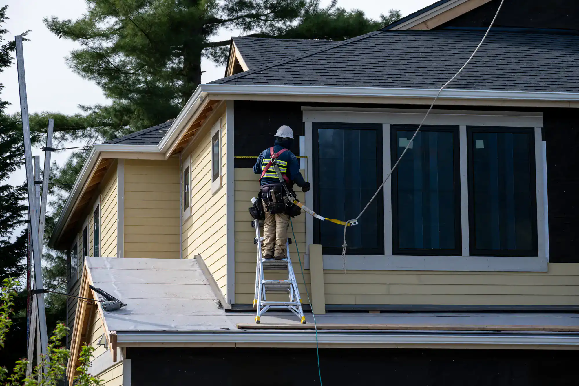 A construction worker wearing safety gear stands on a ladder placed on a sloped roof, working on the exterior of a yellow house with large windows and black trim. Tall trees are visible in the background.