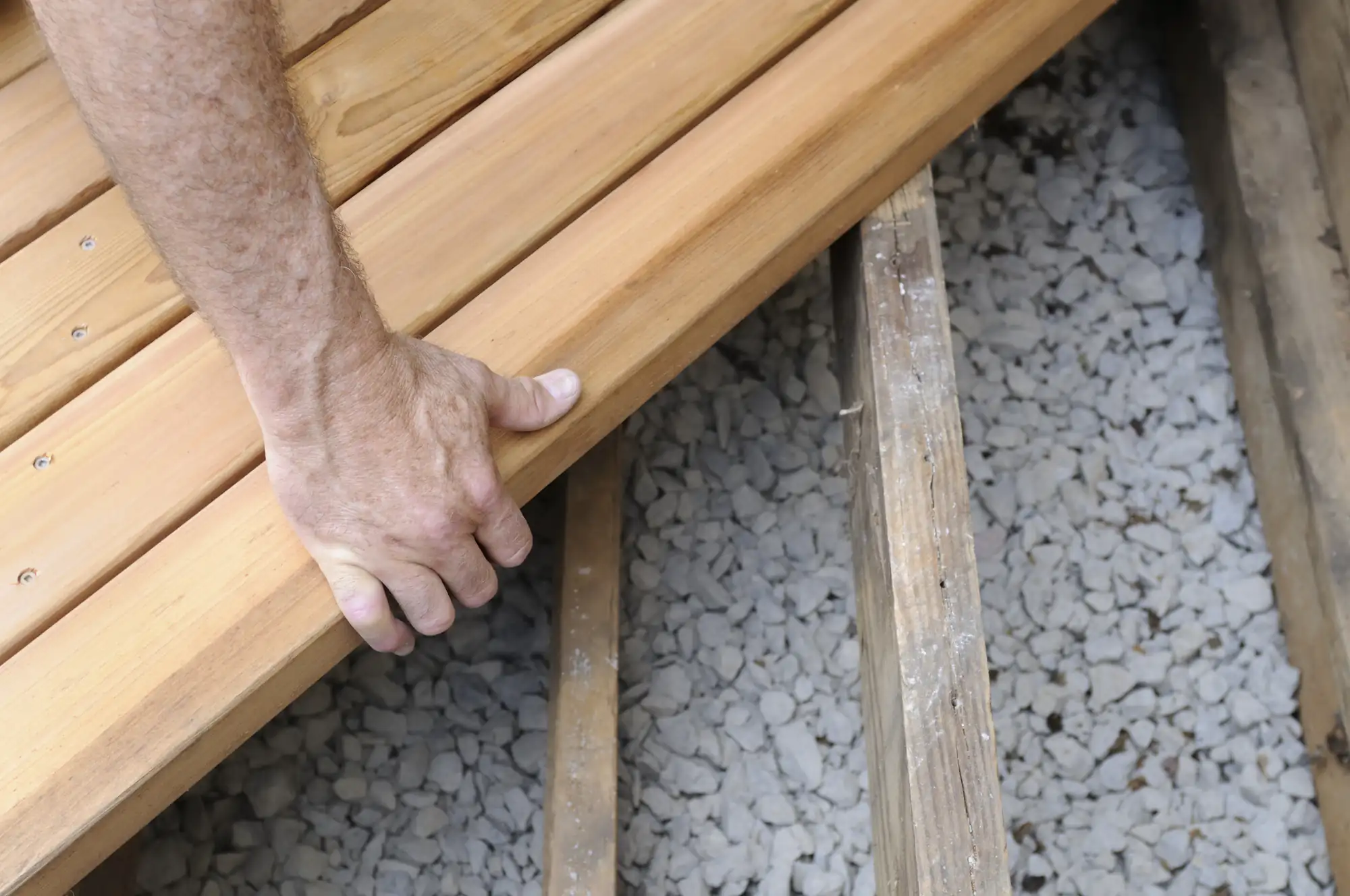 A person&rsquo;s hand is placing or adjusting a wooden plank onto a deck frame above a layer of gravel, suggesting the construction or installation of a wooden deck.