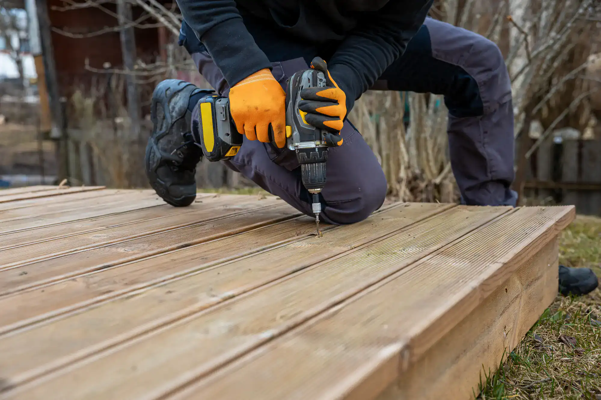 A person wearing orange gloves uses a power drill to drive a screw into a wooden deck while kneeling outdoors.