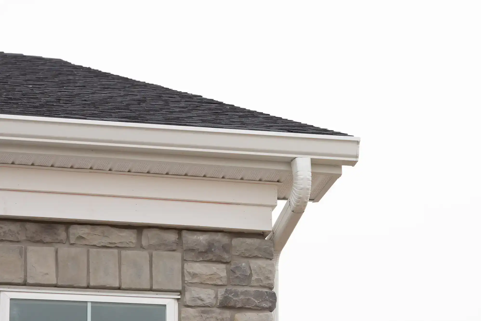 The image shows the corner of a house with stone siding, a shingled roof, and a white rain gutter and downspout against a plain white sky.
