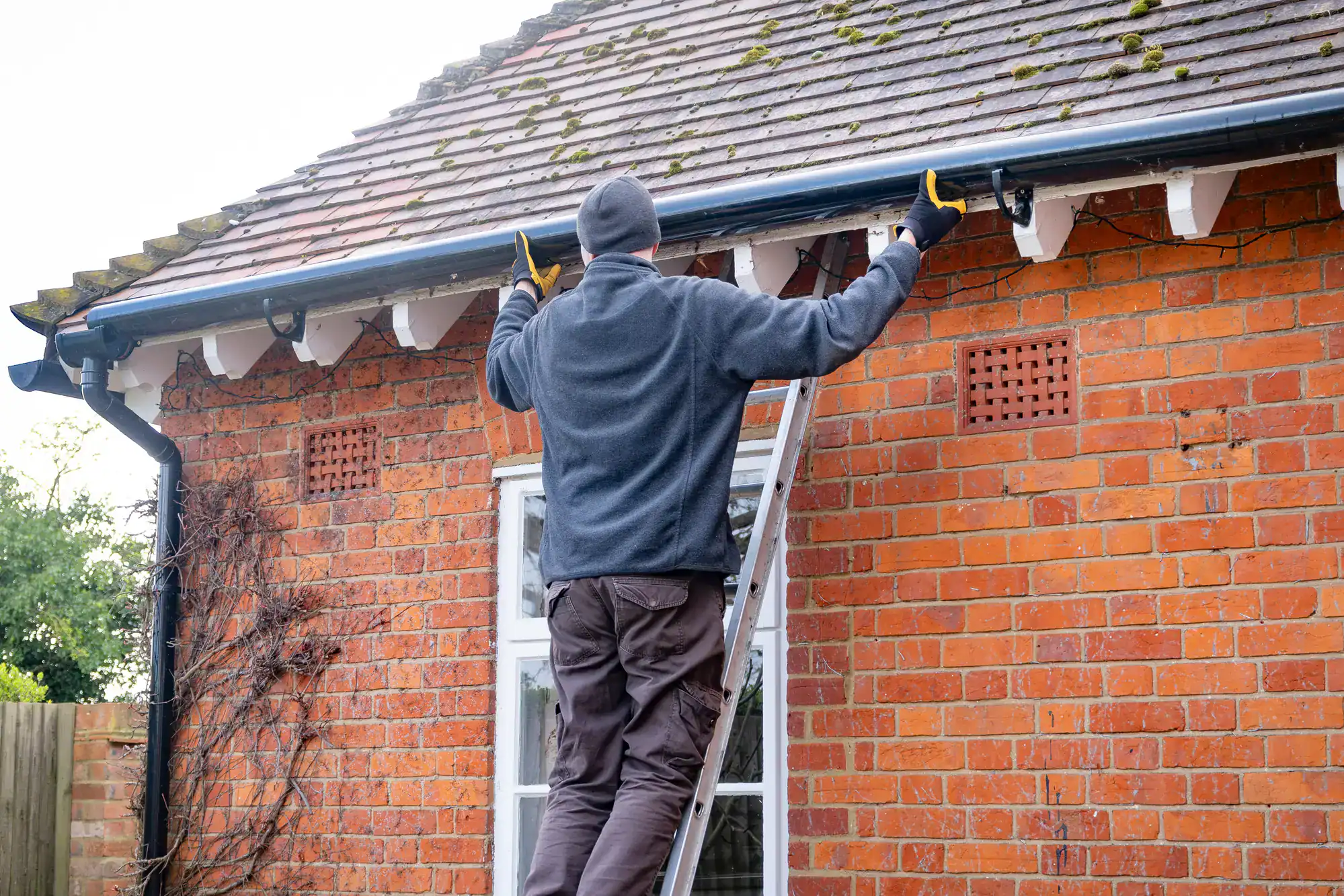 A person standing on a ladder cleans or repairs the gutter of a brick house, wearing a beanie, gloves, and a fleece jacket. The roof has some moss, and the weather appears cloudy.