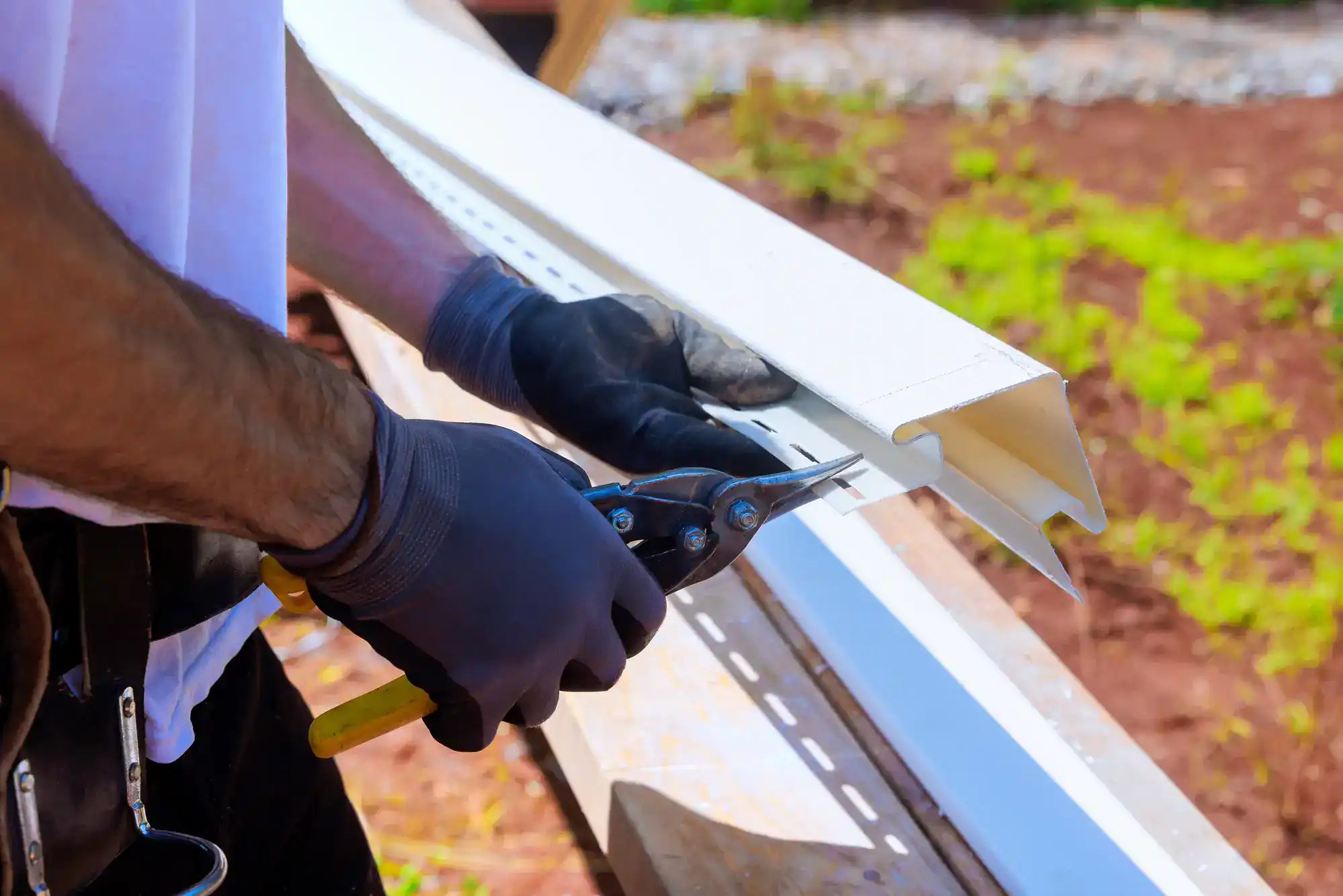 A person wearing black gloves uses tin snips to cut a piece of white metal siding or trim, working outdoors on a sunny day with grass and soil visible in the background.