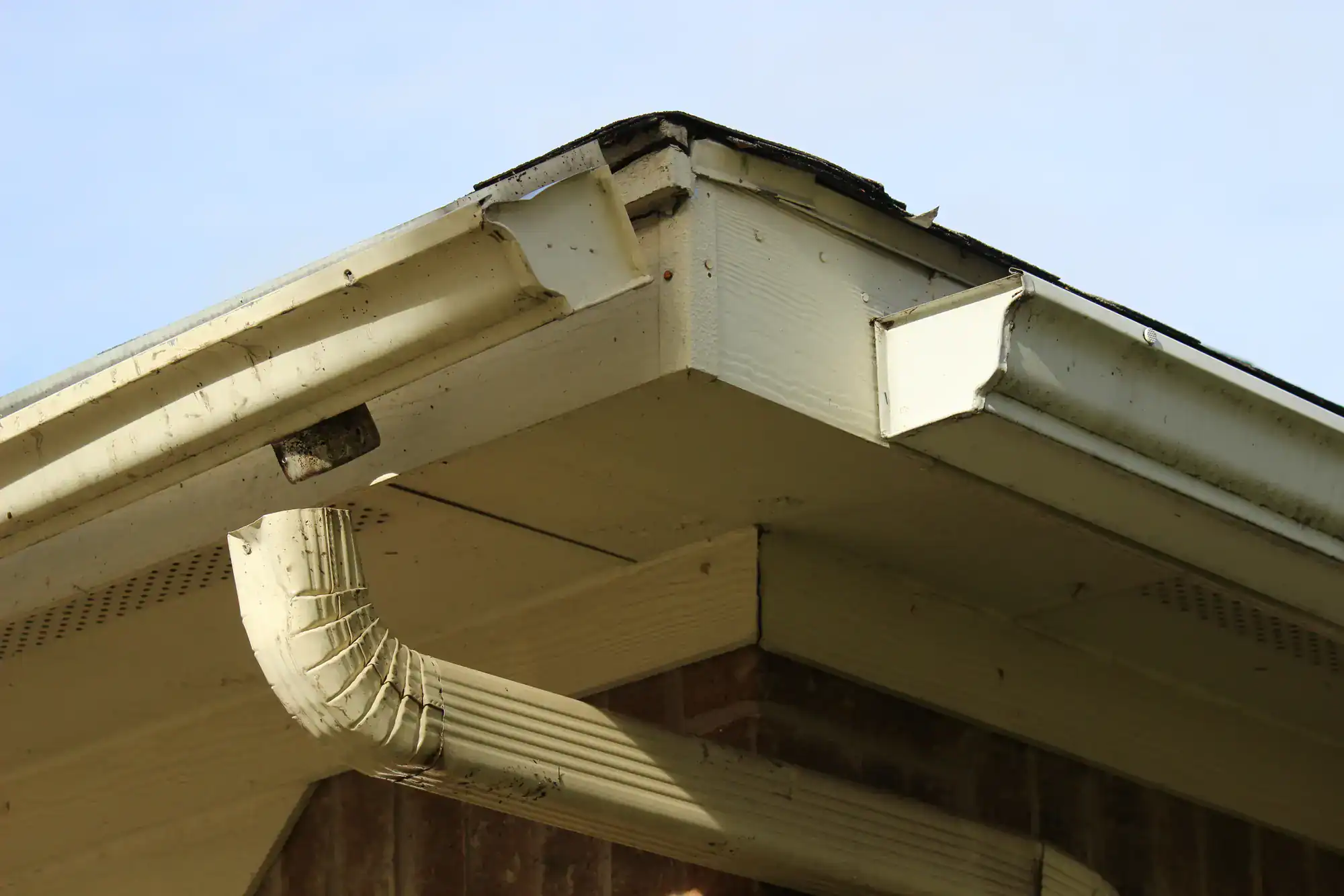 A close-up of a house roof corner with damaged, sagging gutters, peeling paint, and dirty spots. The downspout is attached to the brick wall below. The sky is clear in the background.