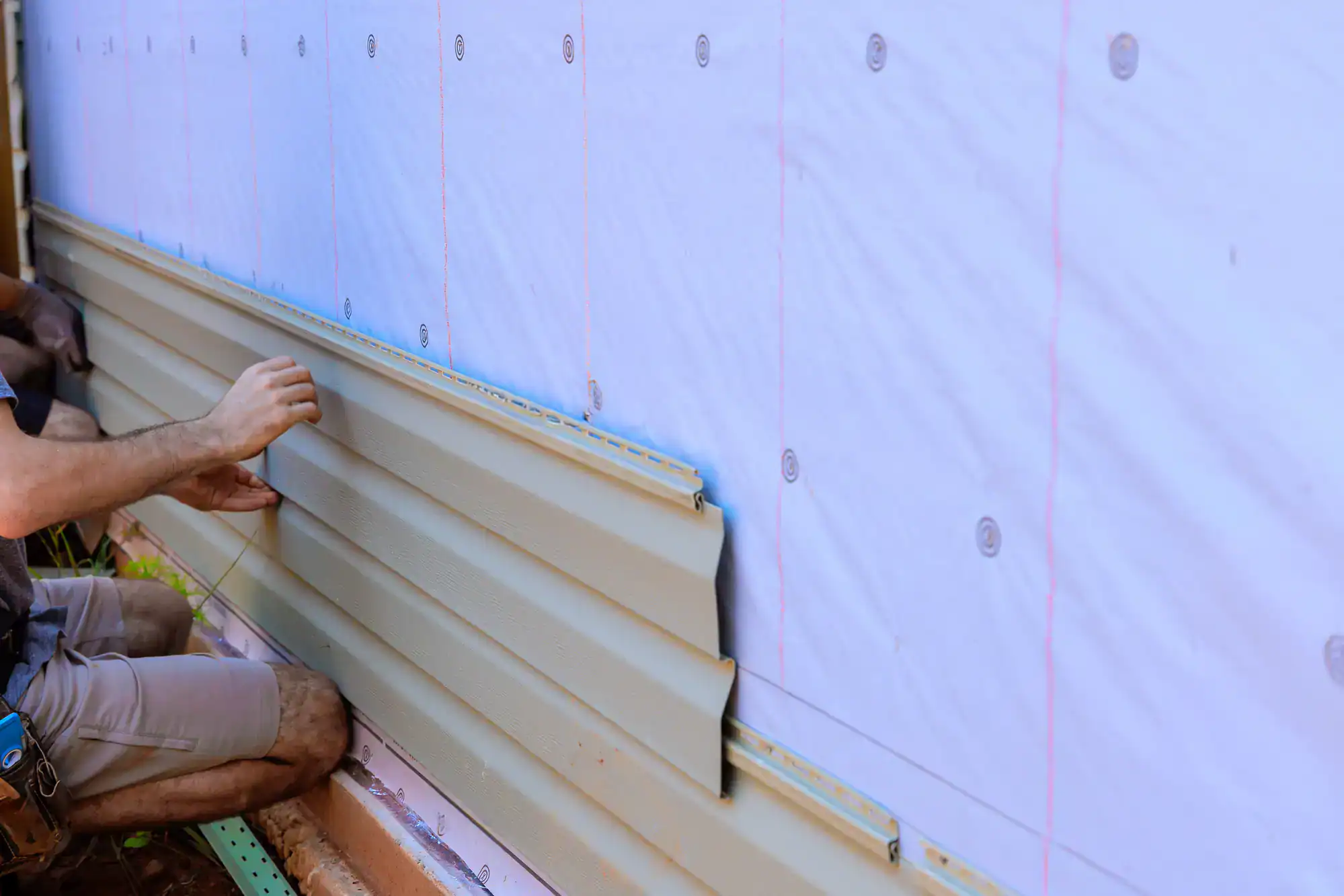 A person installs beige horizontal vinyl siding panels on the exterior wall of a house, which is covered with a white weather-resistant barrier.