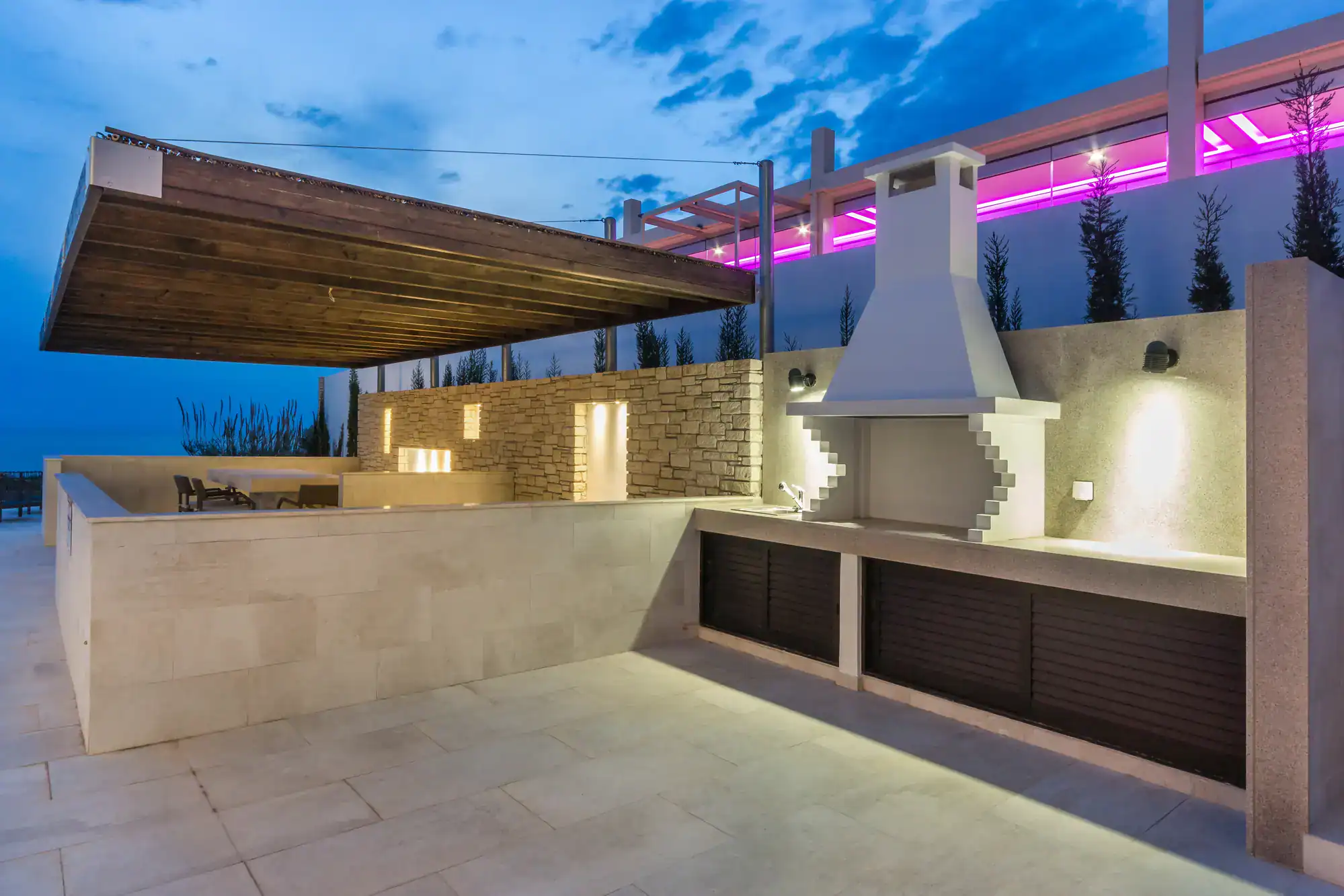 Modern outdoor kitchen with a built-in grill and chimney, stone counter, wooden canopy, and accent lighting. The area is lit with wall lights and purple LED lights, with a seating area in the background at dusk.