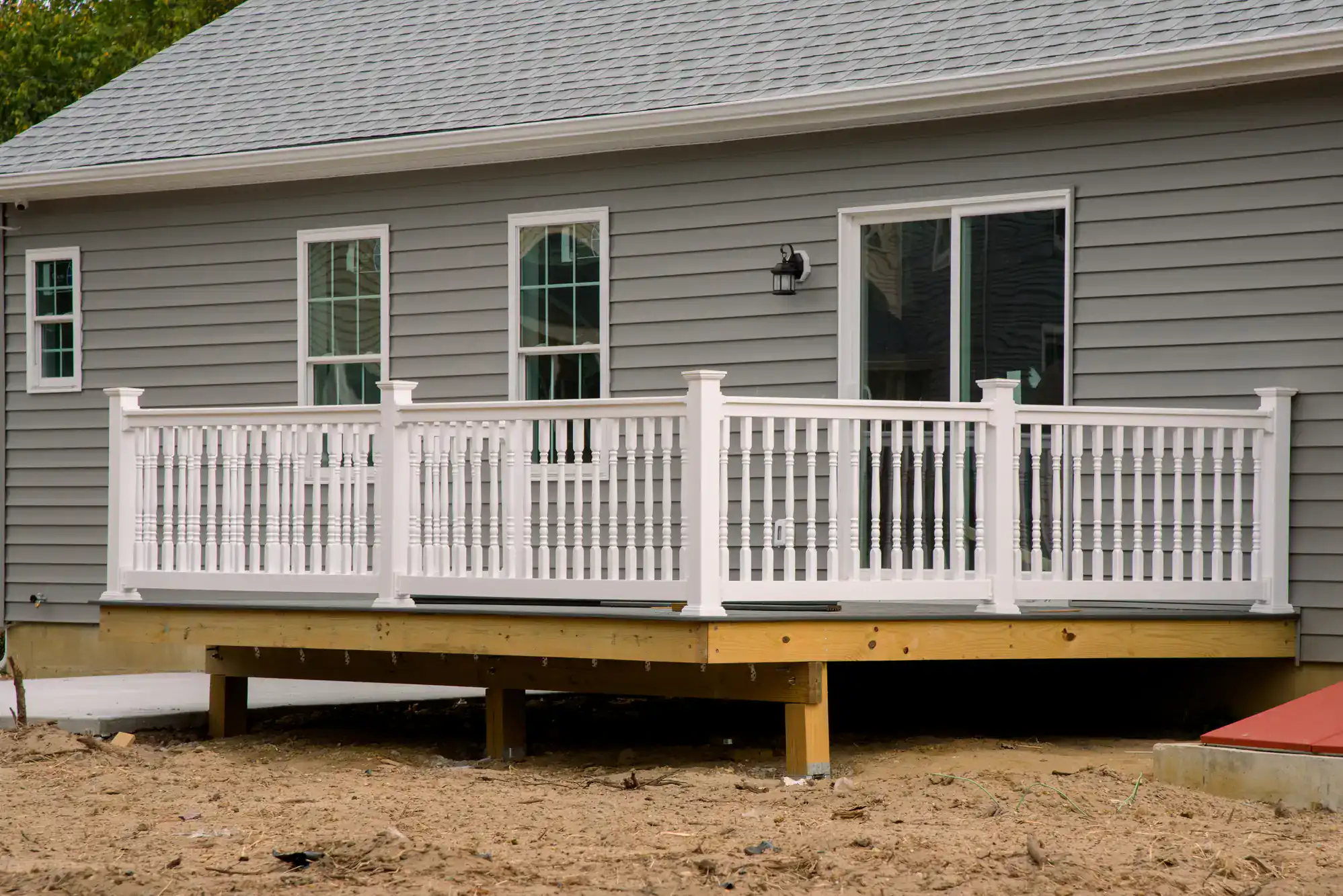 A small, newly built wooden deck with white railings attached to a gray house with sliding glass doors and two windows. The ground below the deck is bare dirt.