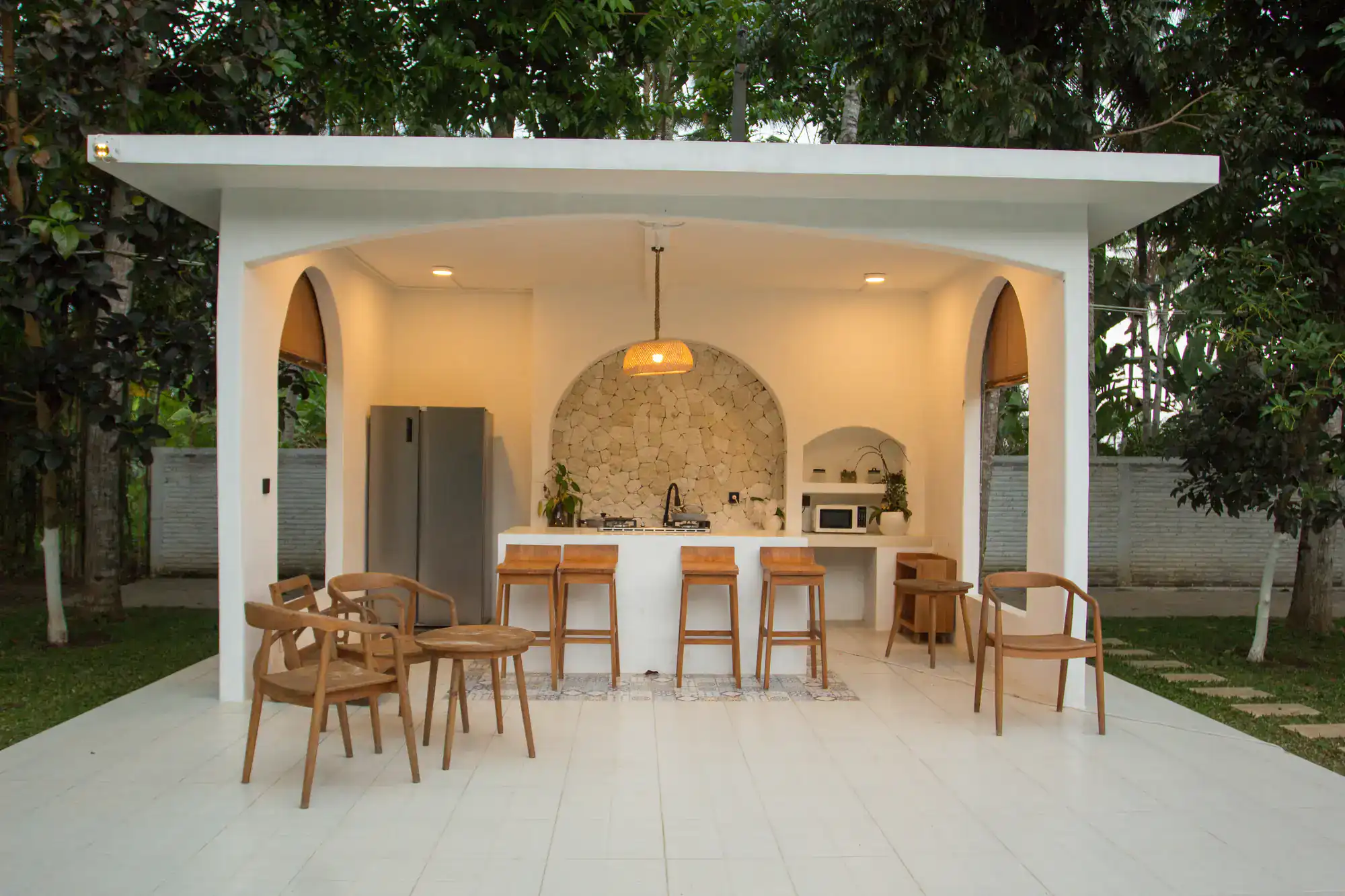 Open-air modern outdoor kitchen with a white countertop, wooden barstools, a refrigerator, microwave, and decorative lighting, surrounded by greenery and trees.
