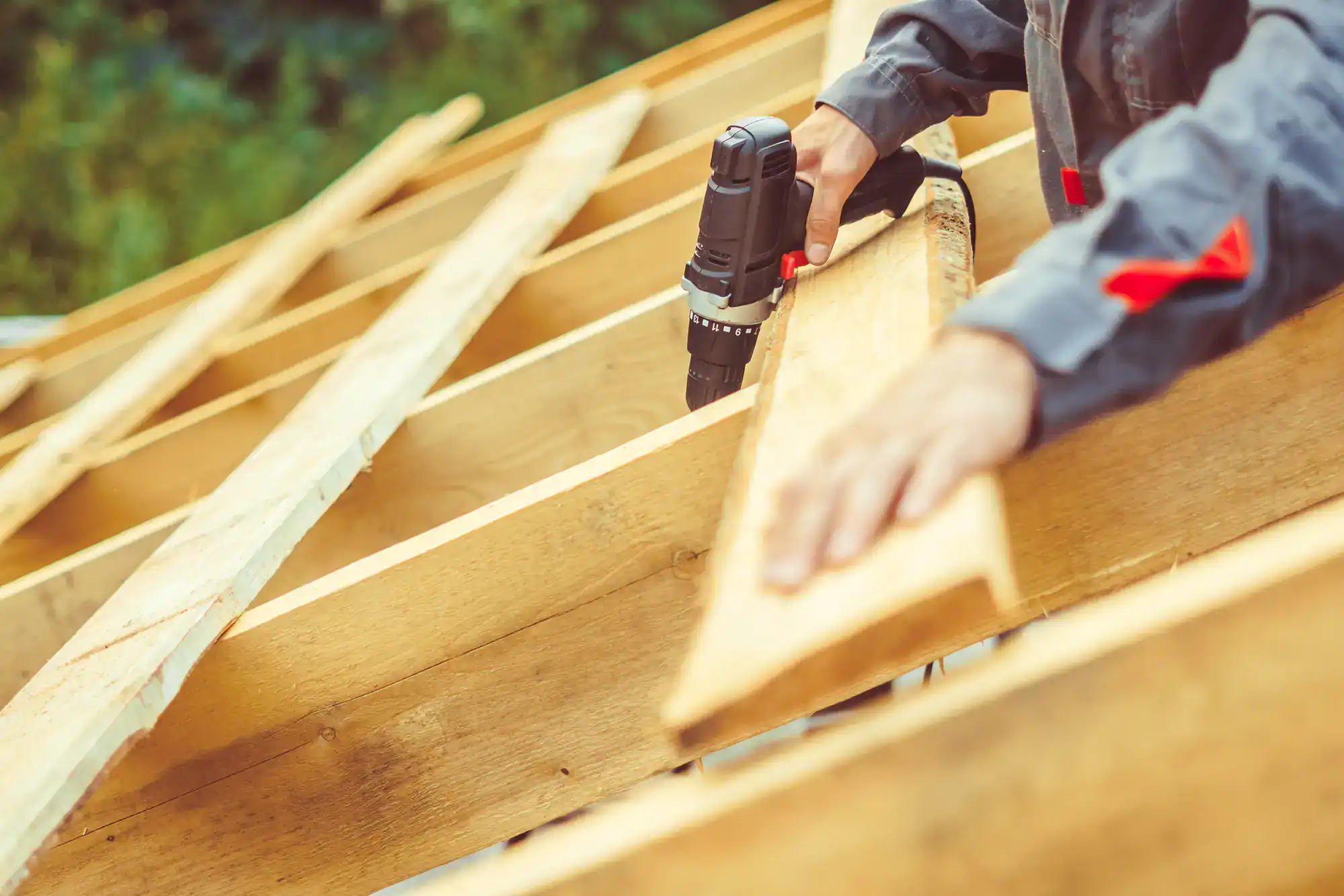 A person wearing a gray work uniform is using a cordless power drill on wooden beams, constructing a roof outdoors with greenery in the background.