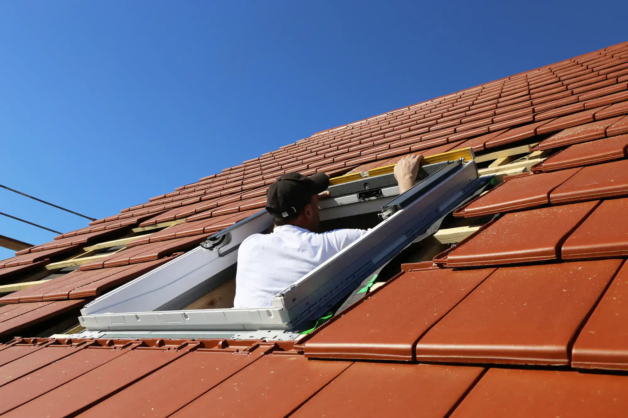A person installing or adjusting a skylight window on a sloped red tiled roof under a clear blue sky. The worker is using tools and wearing a black cap and white shirt.