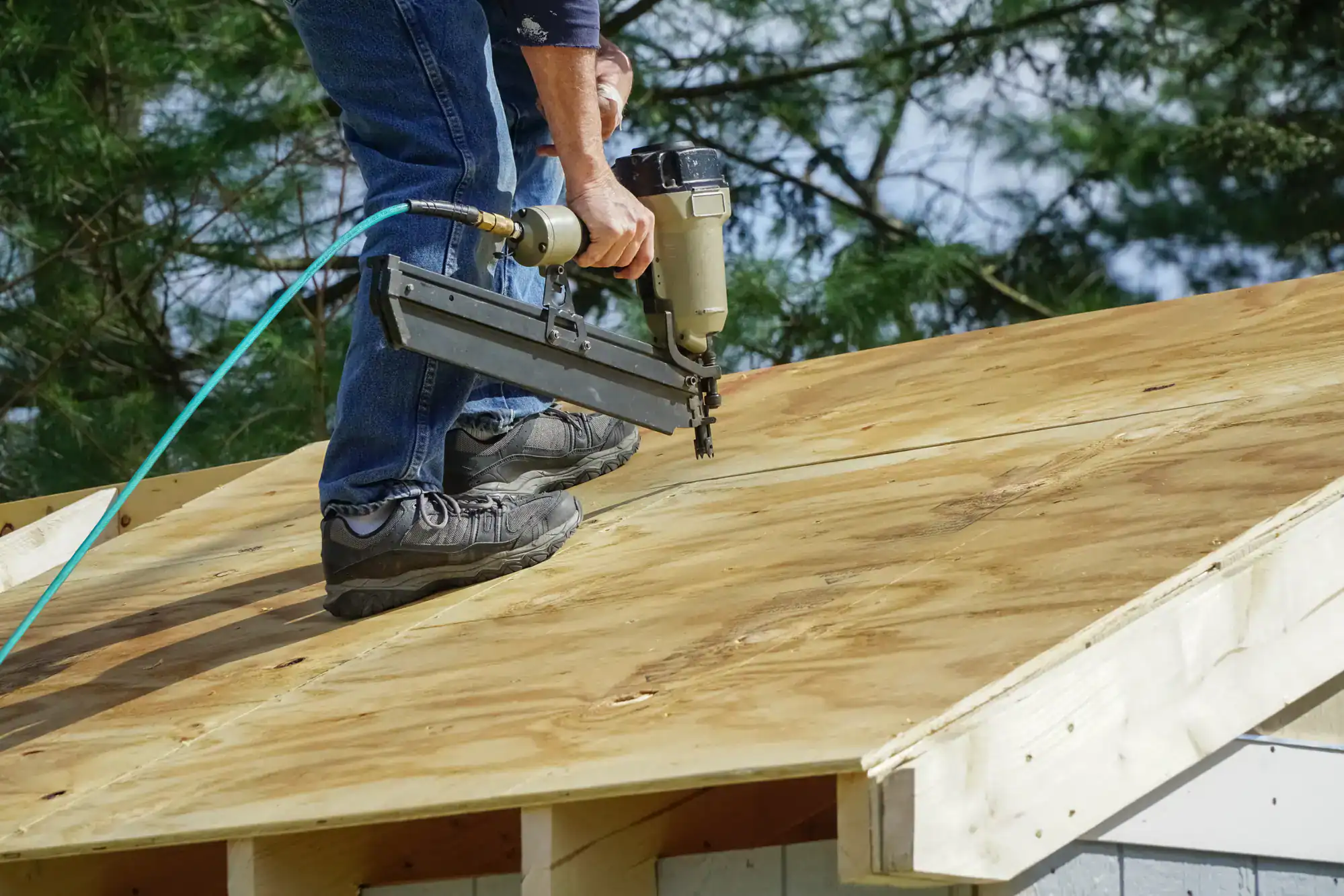A person wearing jeans and work boots uses a nail gun to secure plywood sheets to a roof under construction, with trees visible in the background.