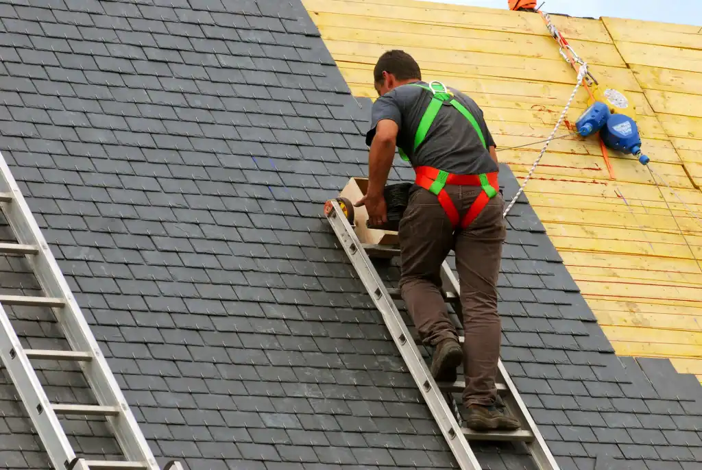A worker wearing a safety harness climbs a ladder on a steep, shingled roof next to a section of exposed wooden planks, with safety equipment visible in the background.