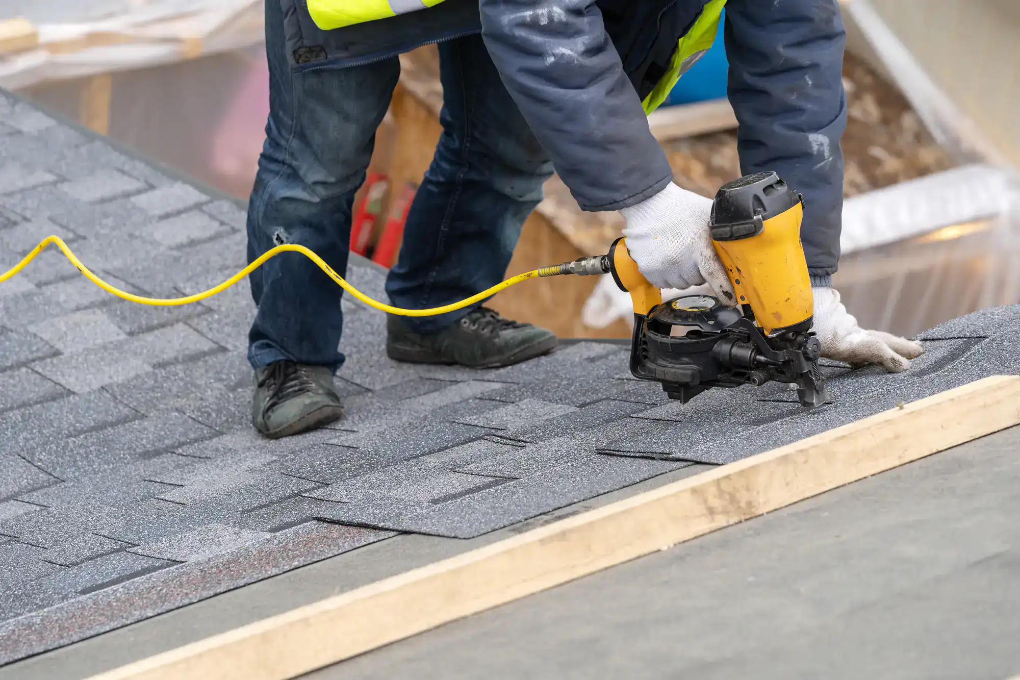 A worker wearing gloves uses a nail gun to install gray asphalt shingles on a sloped roof during construction.